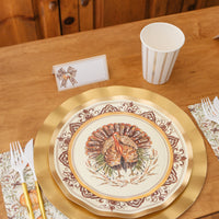 A Sophistiplate LLC Turkey & Gourds: Thanksgiving Table for 16 place setting featuring a decorative turkey heavy duty paper plate on a gold charger, a striped paper cup, a place card with a bow, and a napkin decorated with gourds.