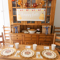 A Thanksgiving table setting featuring the Turkey & Gourds: Thanksgiving Table for 16 set with heavy-duty paper plates, gold chargers, and cups, with a happy fall banner hung on the hutch behind the table.
