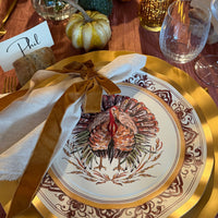 A Sophistiplate LLC Turkey & Gourds: Thanksgiving Table for 16 setting featuring a white turkey-illustrated heavy duty paper plate layered over a gold charger, complete with a cloth napkin, velvet ribbon tie, and a place card labeled Phil.