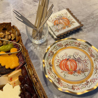 A charcuterie board next to Turkey & Gourds: Thanksgiving Table for 16 decorative plates and pumpkin-patterned napkins, with a glass holding wooden appetizer forks.