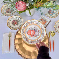 A hand sets the Sophistiplate Turkey & Gourds Table Setting, featuring a designer paper plate, at a fall-themed table with matching napkin, cutlery, and a floral and succulent centerpiece.