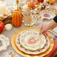 A hand arranges the Sophistiplate Turkey & Gourds Table Setting on an autumn table with Thanksgiving tableware—gold-rimmed plates, candles, glasses, mini pumpkins—plus a fall-patterned cloth and napkin.