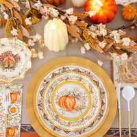 Create a warm and inviting Thanksgiving tablescape with the Turkey & Gourds Table Setting by Sophistiplate, featuring pumpkin-themed designer paper plates, gold chargers, faux pumpkins, candles, a floral garland, and vintage turkey napkins. White and gold flatware is arranged beside each place setting atop an orange tablecloth for a festive autumn look.