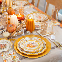 The Turkey & Gourds Table Setting by Sophistiplate features designer plates, gold chargers, candles, decorative pumpkins, glassware, and cutlery on a floral tablecloth with a festive fall centerpiece.